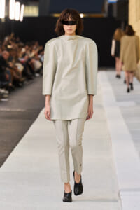 Model walking a runway in a pale beige outfit with exaggerated shoulder volume, matching slim pants, black visor-style eyewear, and black shoes.