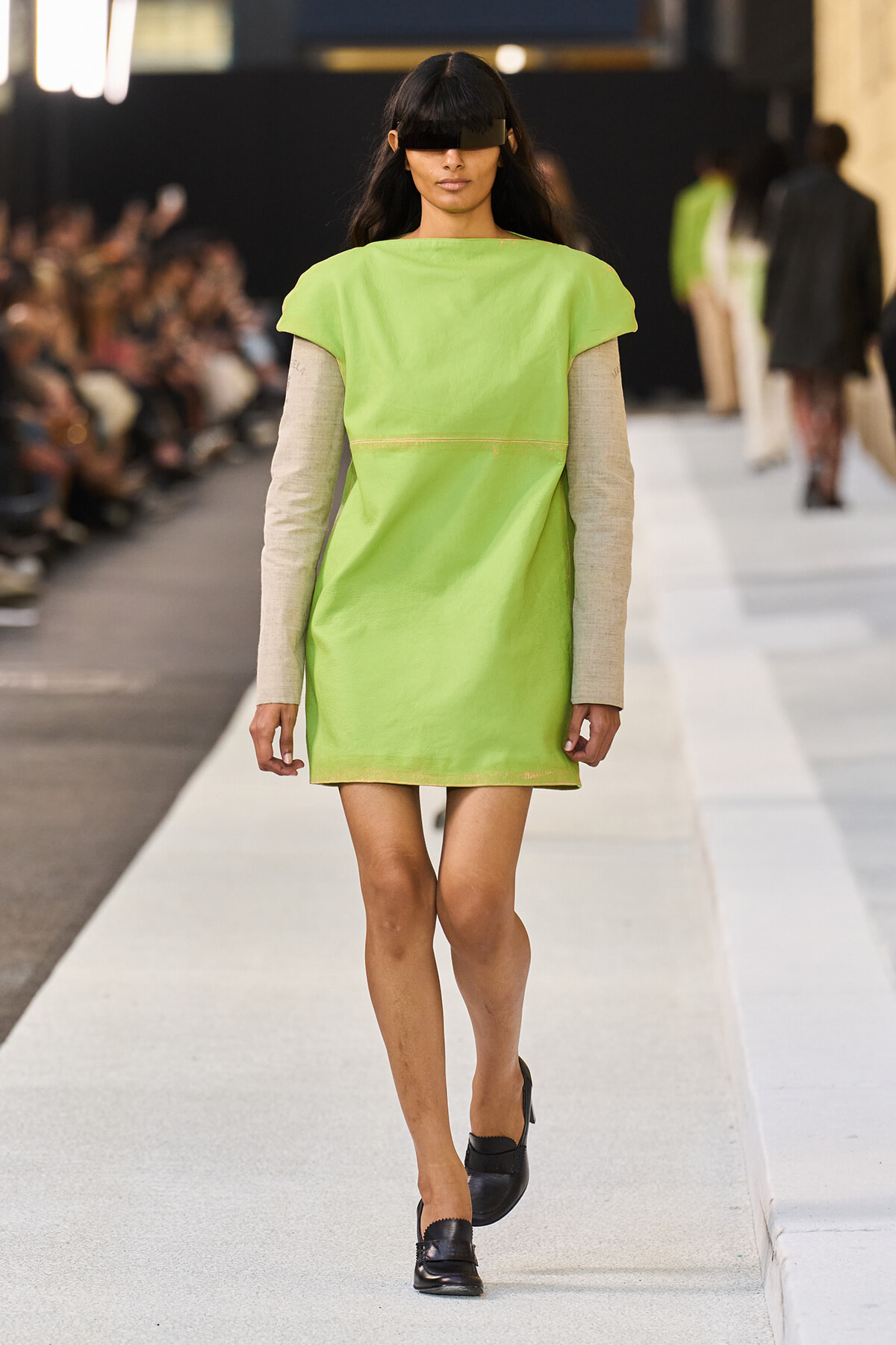 Model walks the runway in a lime-green shift dress over beige long sleeves, with a black rectangular eye shield.