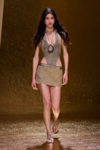 Model walking a runway in a metallic gold halter bodysuit with a pendant necklace and a matching sequin mini skirt, gold sandals, against a gold backdrop.