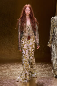 Model walking the runway in a sheer gold lace blouse and gold-leaf printed wide-leg pants with a large stone belt buckle.