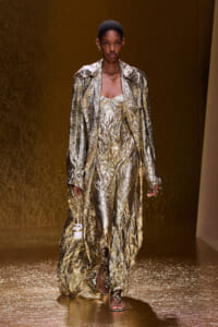 Model on a gold-toned runway wearing a crinkled metallic gold suit and long coat, holding a small white bottle in one hand.