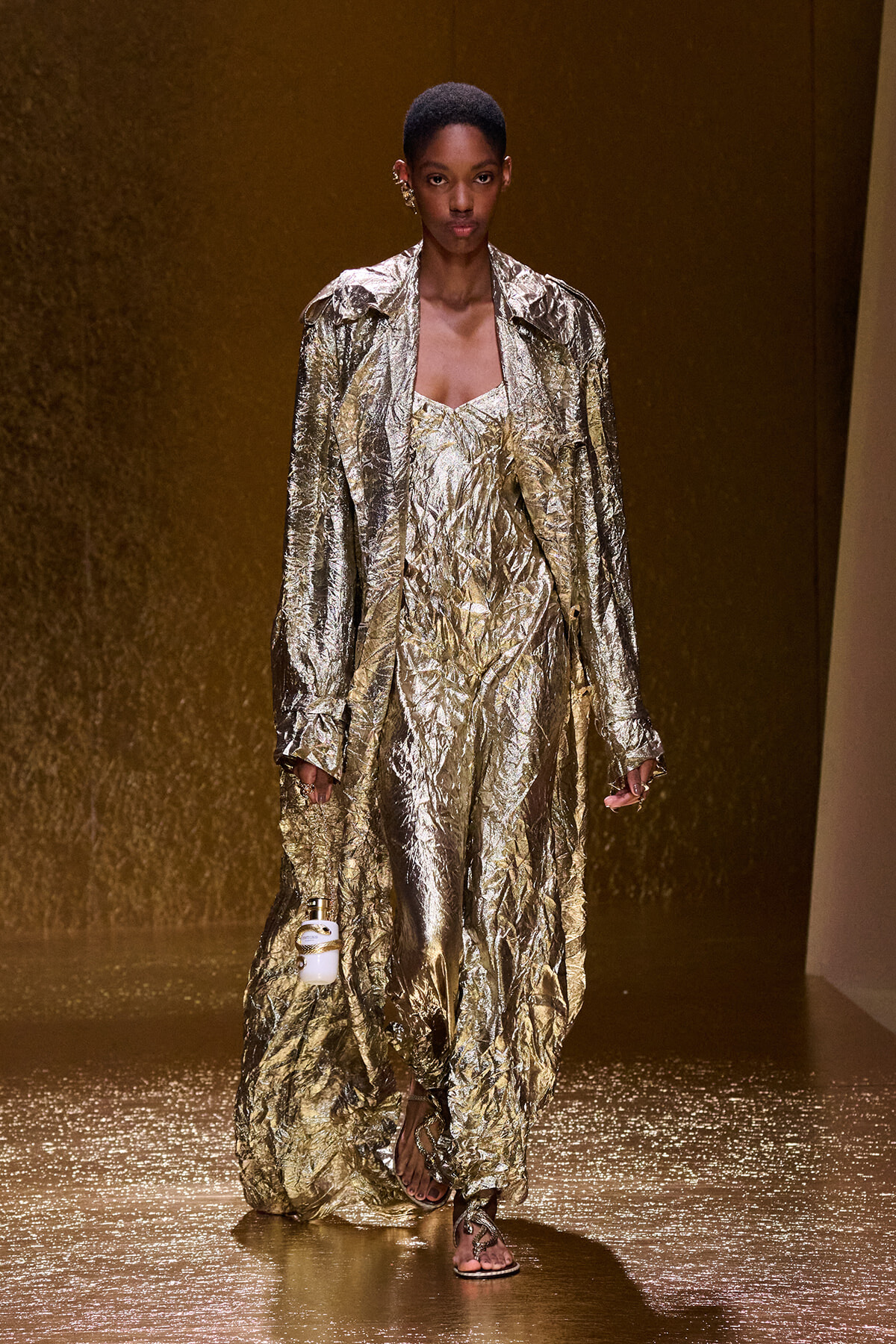 Model on a gold-toned runway wearing a crinkled metallic gold suit and long coat, holding a small white bottle in one hand.