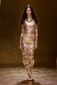 Model walking the runway in a shiny gold metallic long-sleeve gown with a statement collar necklace.