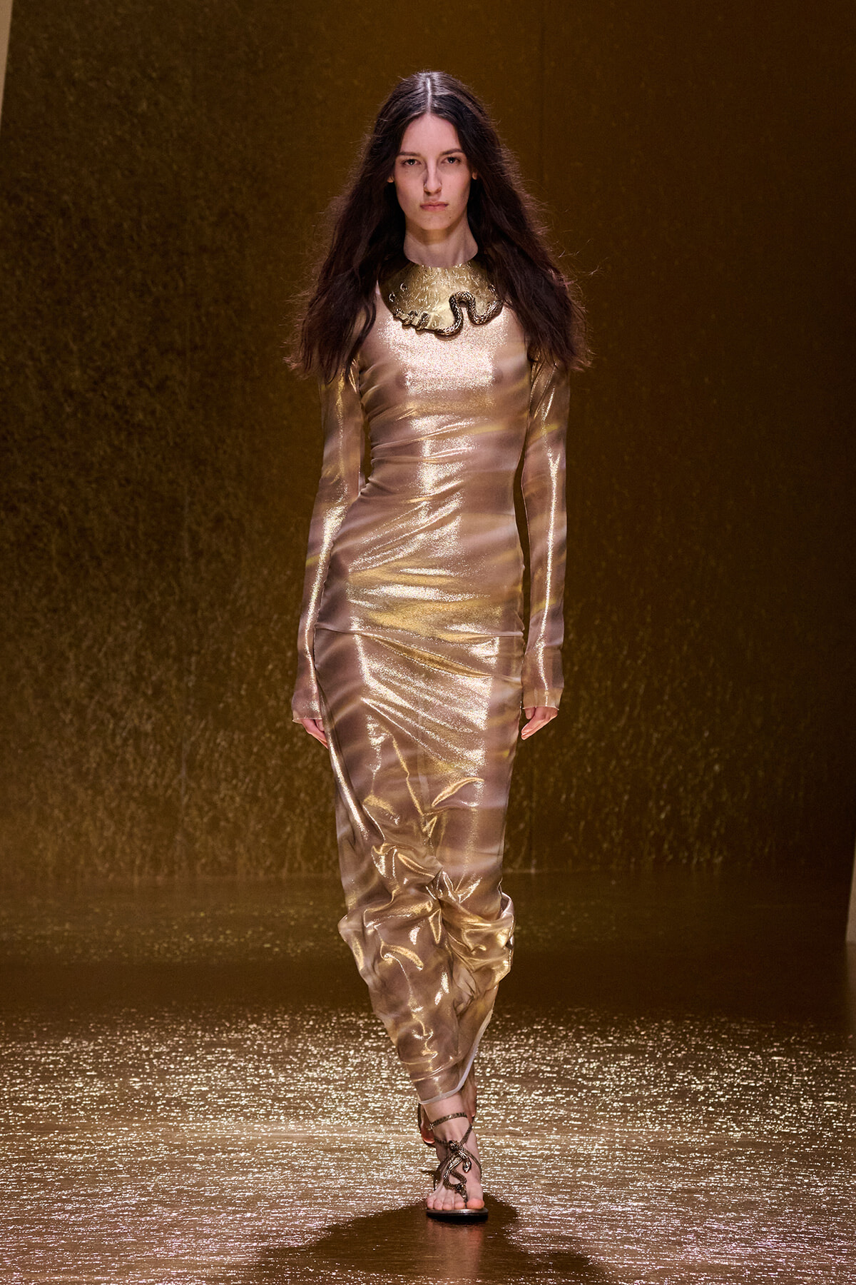 Model walking the runway in a shiny gold metallic long-sleeve gown with a statement collar necklace.