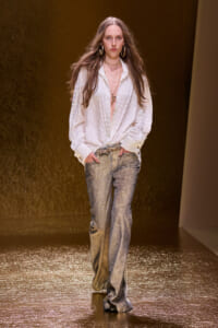 Runway model in a white textured blouse with shimmering silver trousers and gold jewelry on a golden runway stage.