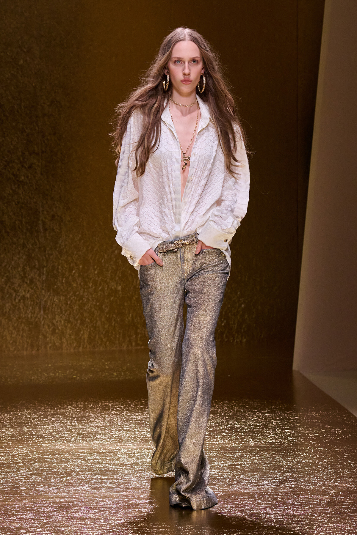 Runway model in a white textured blouse with shimmering silver trousers and gold jewelry on a golden runway stage.