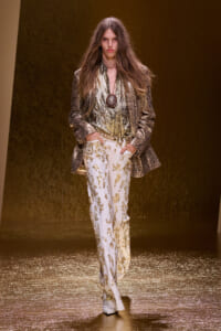 Model walking the runway in a metallic gold blazer and shirt with white embroidered pants, accessorized with a large circular pendant necklace on a glittery floor.