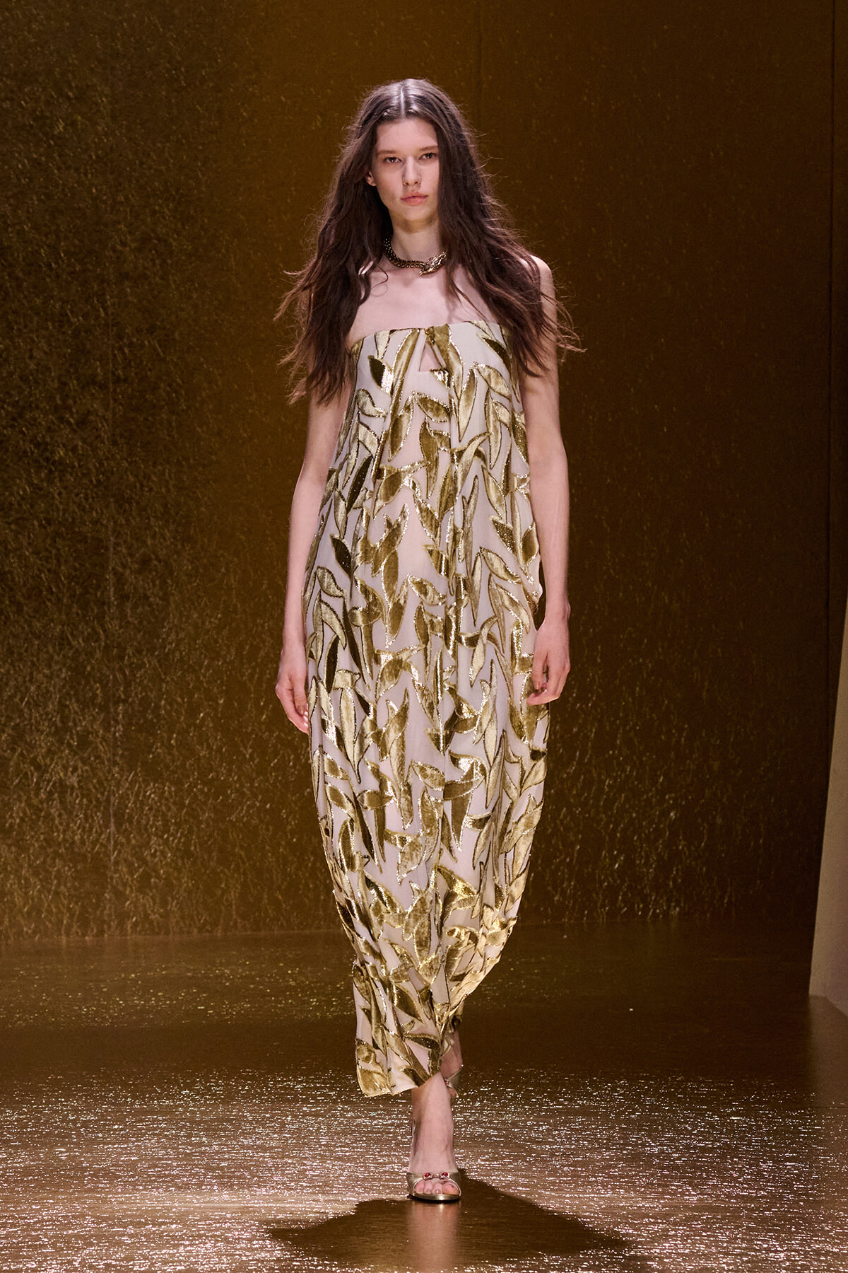 Model walking a runway in a strapless beige gown with gold leaf appliqué pattern.