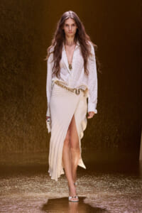 Model walking a fashion show in a white textured blouse, cream pleated skirt with a gold chain belt and matching clutch.