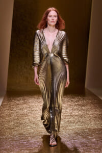Model on a runway wearing a gold metallic pleated gown with a deep V neckline.