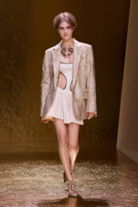 Model walks the runway in a metallic gold blazer over a white cutout top and pleated mini skirt, with a chunky snake necklace and snakeskin heels.