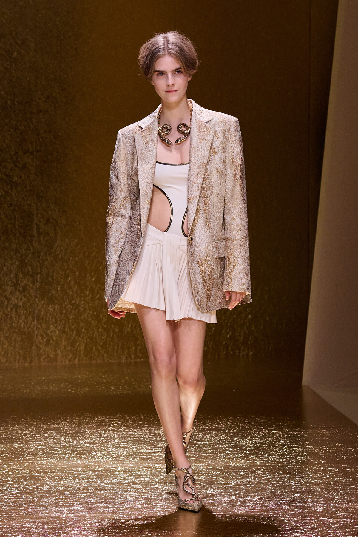 Model walks the runway in a metallic gold blazer over a white cutout top and pleated mini skirt, with a chunky snake necklace and snakeskin heels.