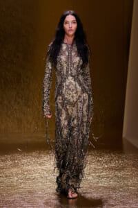 Model walks a runway in a full-length metallic lace gown with black fringe detailing at the sleeves and hem.