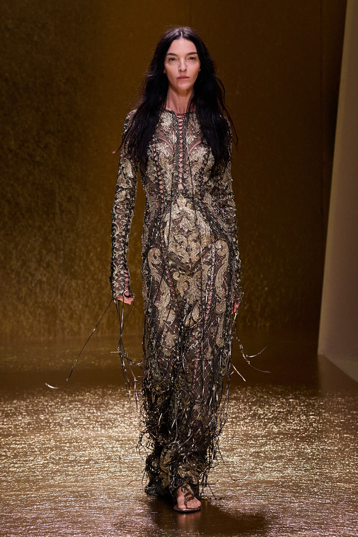Model walks a runway in a full-length metallic lace gown with black fringe detailing at the sleeves and hem.