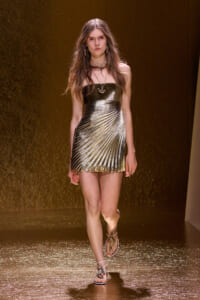 Model walks a fashion show wearing a gold metallic strapless mini dress with diagonal pleats and strappy sandals.