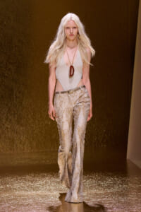 Model walks the runway in a white plunging bodysuit and metallic gold patterned flared pants, with a long red pendant necklace and blonde hair.