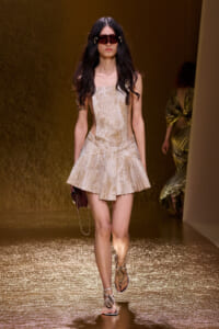 Model walks the runway in a beige textured mini dress with a flared skirt, paired with sandals and a small burgundy clutch, wearing oversized burgundy sunglasses.