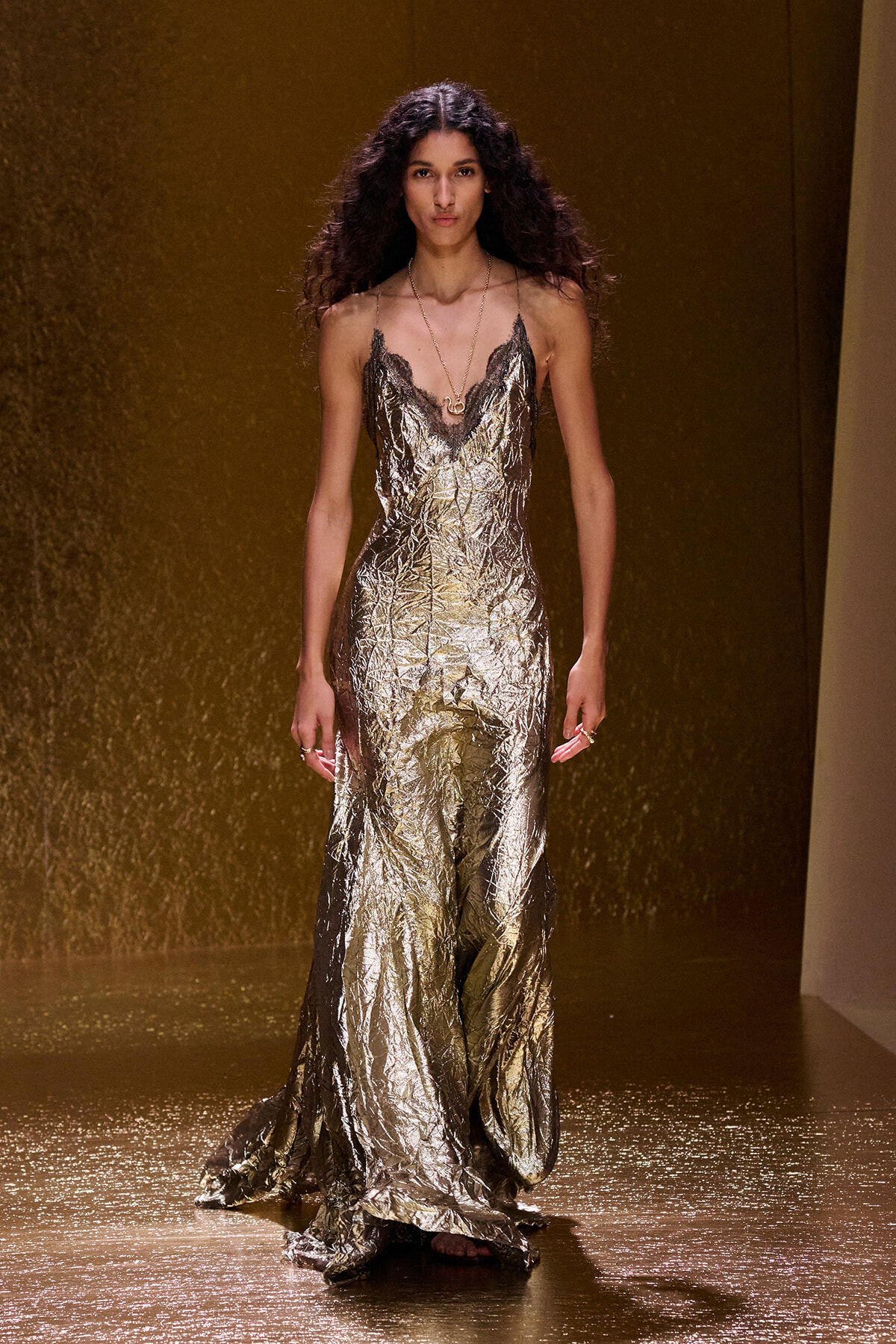 Female model walking a shimmering gold, crinkled-metallic gown with lace-trim neckline on a fashion runway.