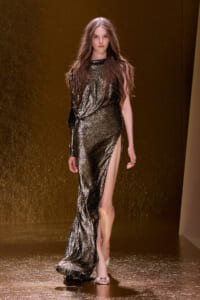 Model walks the runway in a shimmering gold sequined gown with a high slit, neutral expression, long brown hair.