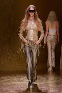 Model walking a fashion runway in a metallic mesh top with fringe sleeves and shimmering wide-leg pants, wearing oversized futuristic sunglasses.