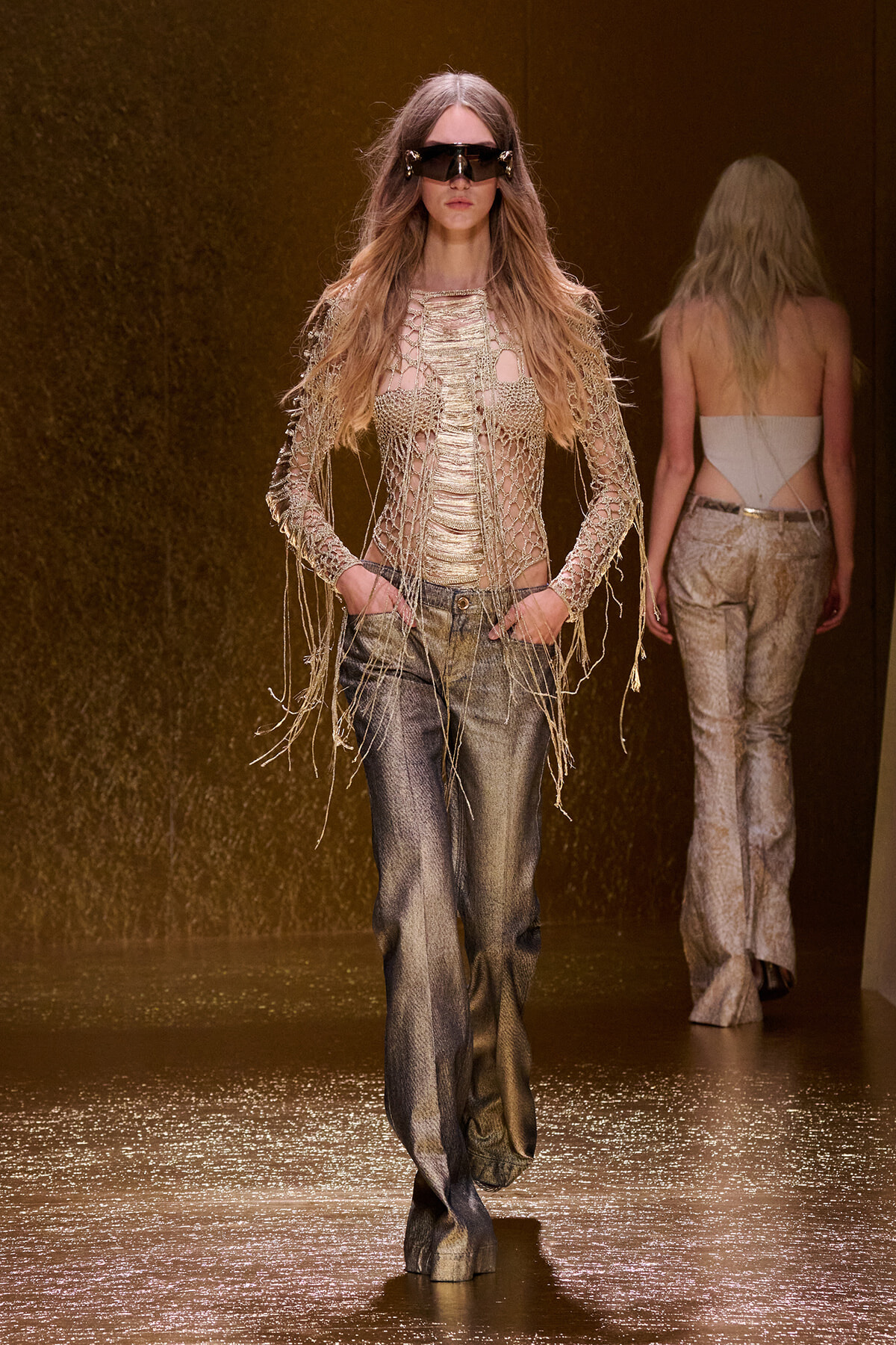 Model walking a fashion runway in a metallic mesh top with fringe sleeves and shimmering wide-leg pants, wearing oversized futuristic sunglasses.