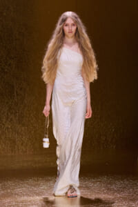 Model walking a runway in a long white textured one-shoulder gown, holding a small white clasped purse on a chain with gold sandals.