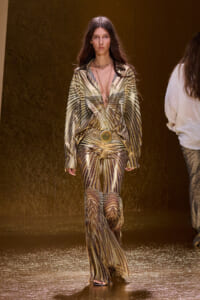 Model walks the runway in a metallic gold, palm-leaf patterned outfit with a plunging neckline and wide belt.