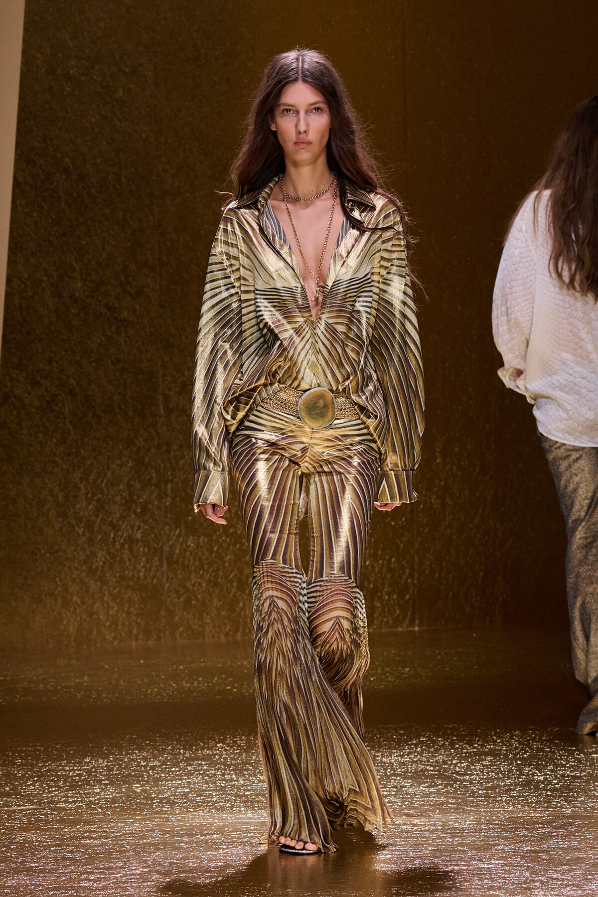 Model walks the runway in a metallic gold, palm-leaf patterned outfit with a plunging neckline and wide belt.
