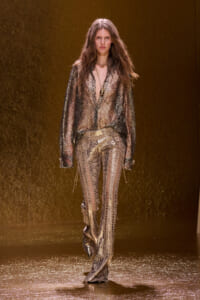 Model walks the runway in a metallic gold snakeskin outfit: sheer lace blouse and matching patterned pants.