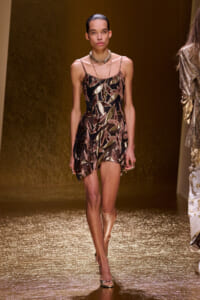 Model walking the runway in a black mini dress with bronze-gold leaf appliqués and layered chains on a glittering gold catwalk.