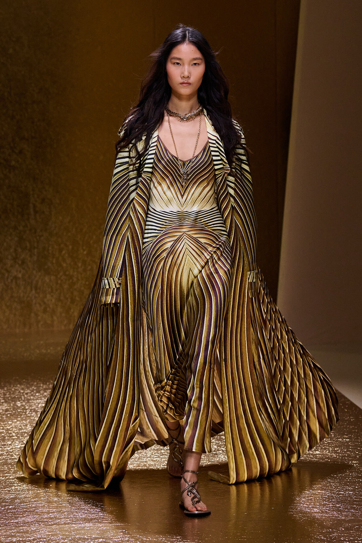 Female model walking a metallic gold-brown striped gown with a long flowing cape on a runway runway show.