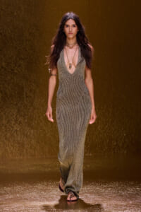Runway model in a shimmering gold halter-neck gown with a deep V, layered necklaces, walking on a glossy gold runway.
