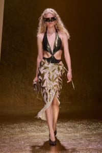 Model walking the runway in a metallic black halter top and gold leaf-patterned skirt, with oversized futuristic sunglasses and a clutch nearby.