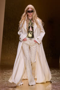 Model on a runway wearing a long white coat over metallic gold halter top and white snakeskin trousers, with oversized futuristic sunglasses and flowing blonde hair.