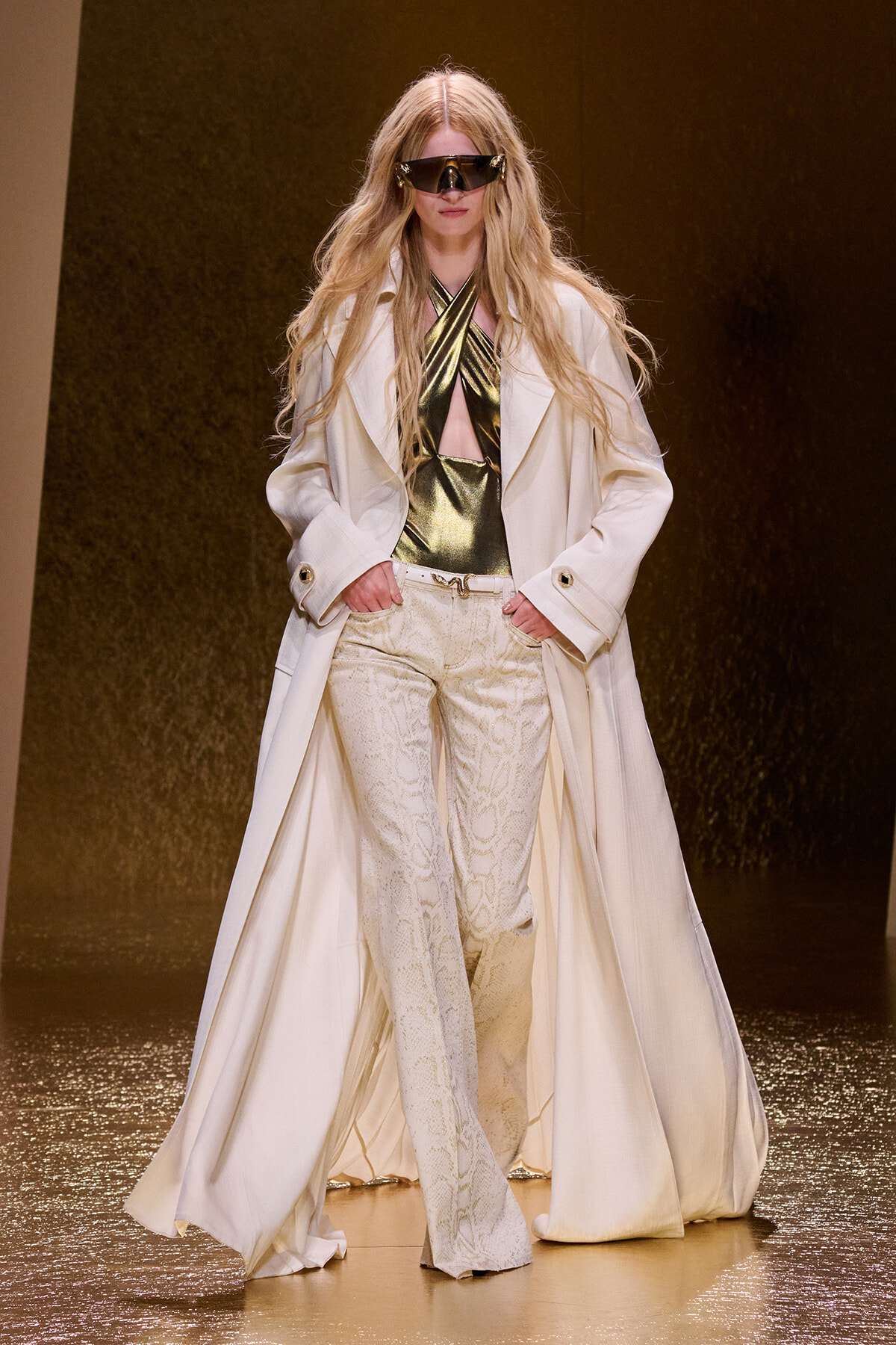 Model on a runway wearing a long white coat over metallic gold halter top and white snakeskin trousers, with oversized futuristic sunglasses and flowing blonde hair.