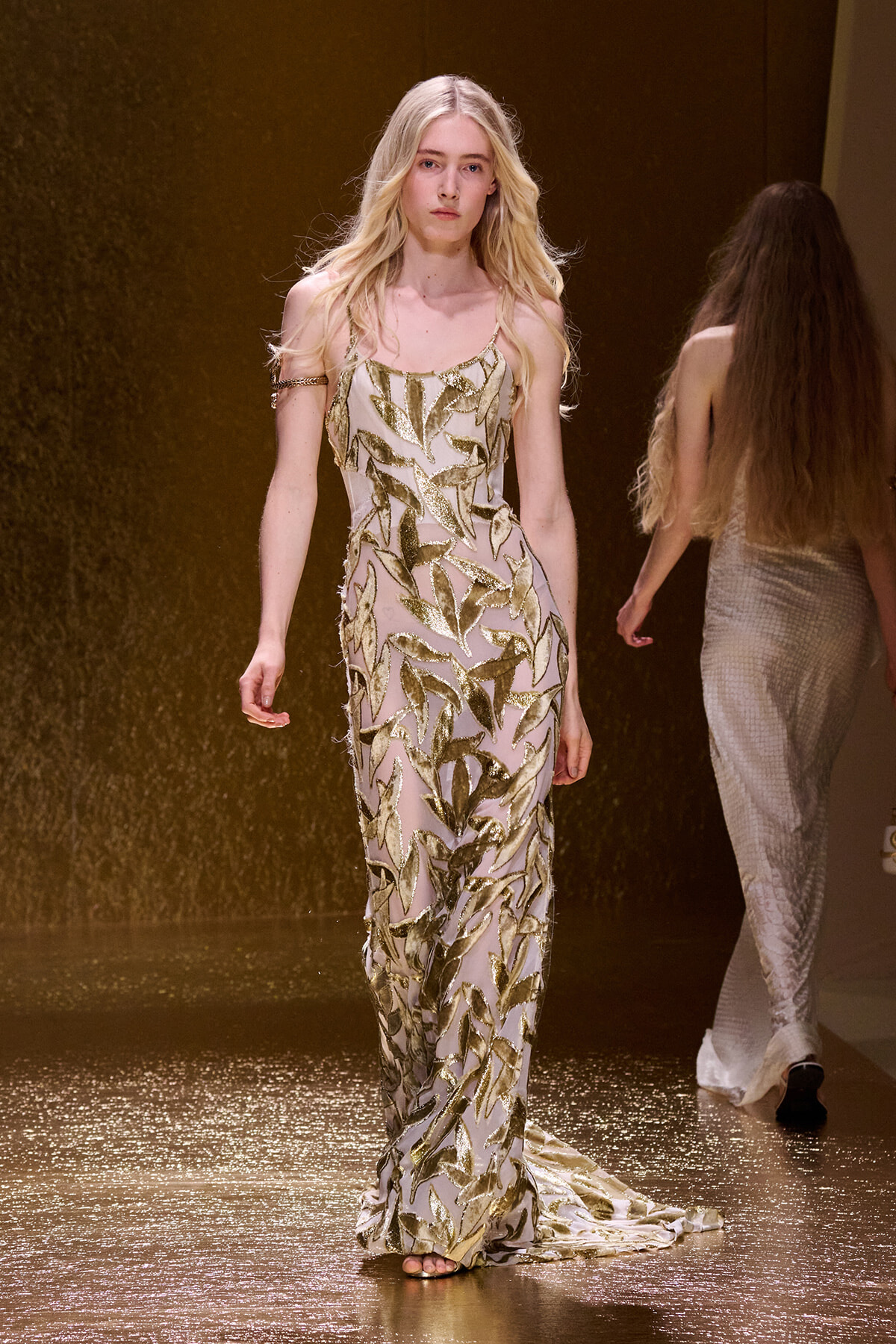 Model walking the runway in a floor-length, sheer gown with gold leaf appliqué patterns.