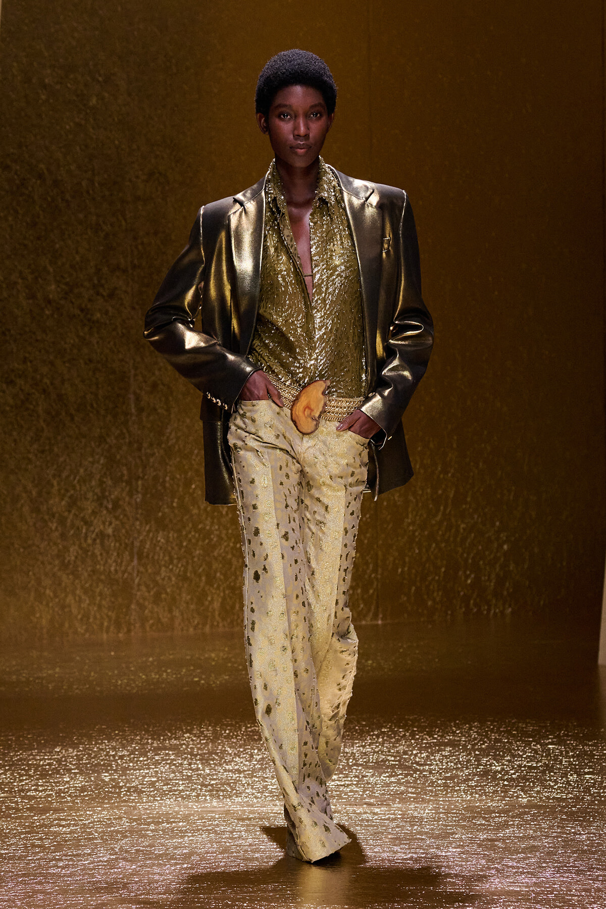 Model walking a gold-themed runway in a metallic gold suit, shimmering shirt, and dotted gold pants, holding an orange accessory at the waist.
