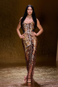 Model walks the runway in a gold sequin gown with a leaf-like pattern, hands on hips.