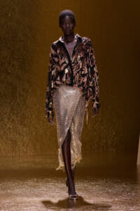 Runway model walking in a metallic gold and black patterned blouse paired with a shimmering gold sequin skirt, against a gold backdrop.
