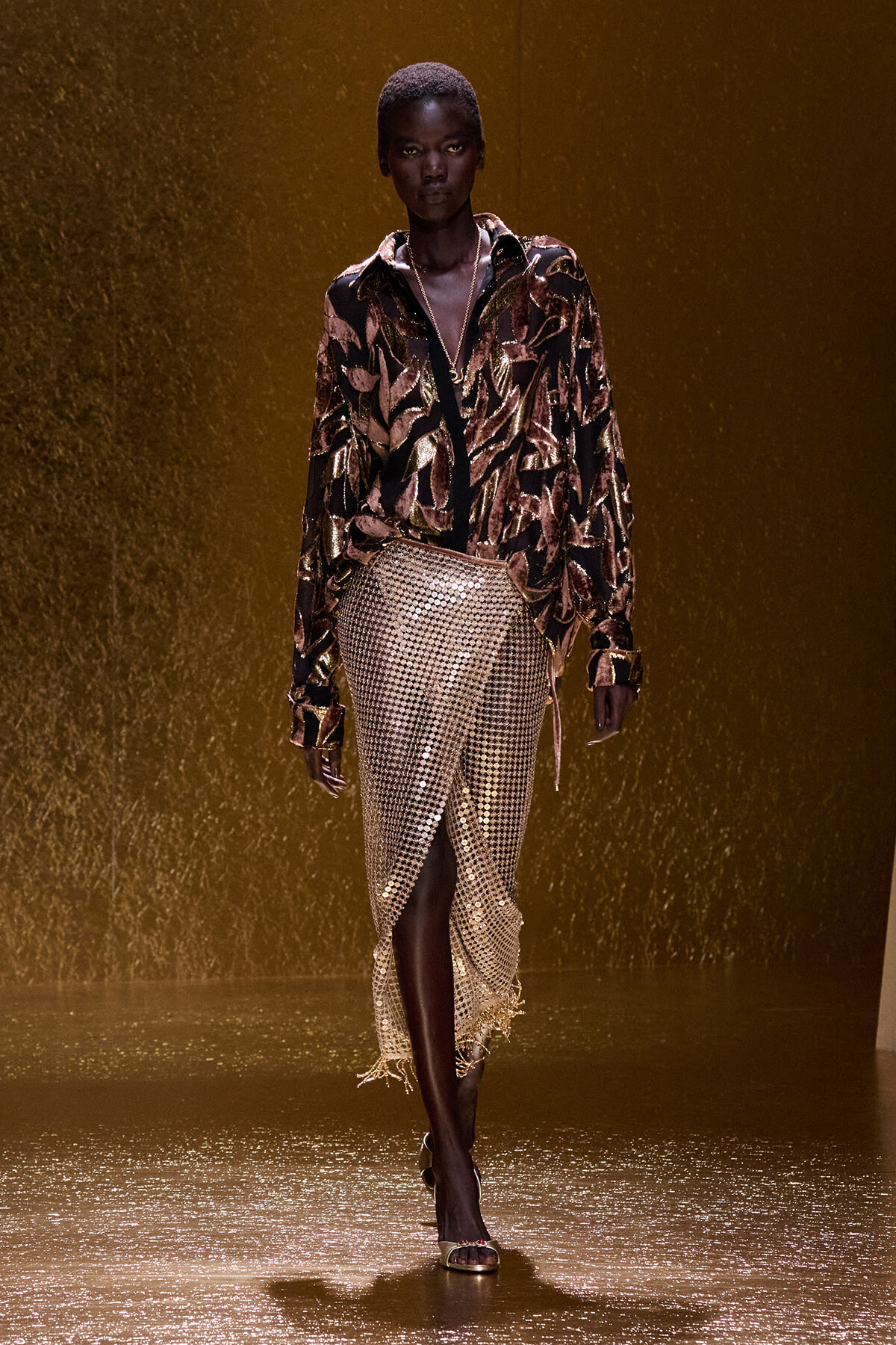 Runway model walking in a metallic gold and black patterned blouse paired with a shimmering gold sequin skirt, against a gold backdrop.