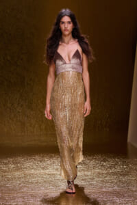 Model walking runway in a shimmering gold evening gown with a metallic V-neck bodice and full sequined skirt.