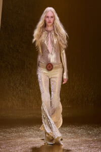 Model on runway wearing a gold metallic snakeskin bodysuit with flared pants and a large circular belt buckle, posing center stage.