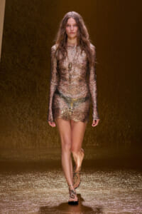 Model walking a fashion runway in a sheer metallic lace mini dress with gold accessories on a gold backdrop while strappy sandals shine under lights.