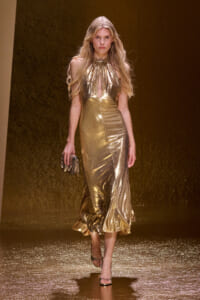 Model walks the runway in a gold metallic halter-neck gown with a keyhole opening, holding a small metallic clutch on a glittering catwalk surface.