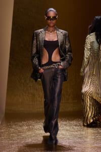 Model wearing a metallic textured blazer over a black cutout bodysuit, gold belt, and futuristic visor on a gold runway.