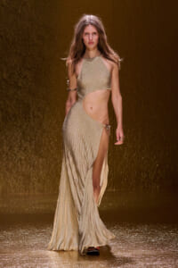 Model walking a fashion runway in a shimmering gold cut-out gown with a high slit and minimalist accessories.