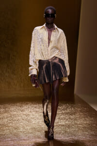 Model on a runway wearing a cream patterned blouse with a metallic pleated brown mini skirt, dark sunglasses, and gold accessories.