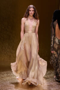 Model walks the runway in a gold, flowing halter gown with a sheer waist and full skirt against a shimmering gold backdrop from the fashion show.