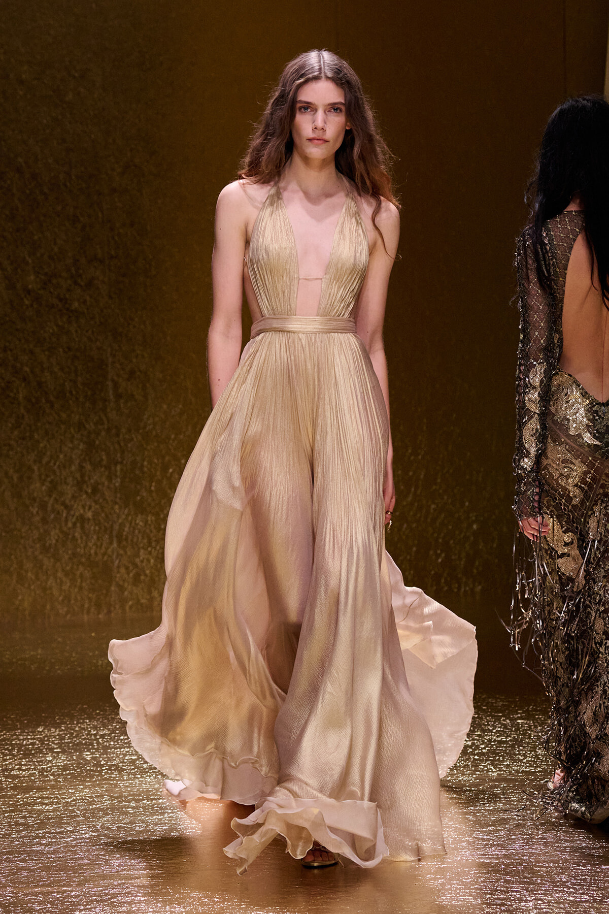 Model walks the runway in a gold, flowing halter gown with a sheer waist and full skirt against a shimmering gold backdrop from the fashion show.