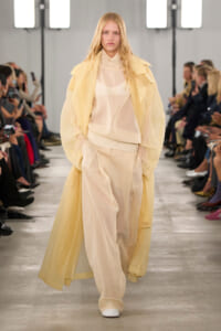 Model walking a fashion show in a sheer cream blouse and wide pale pants under a long pale-yellow coat, runway audience on both sides.